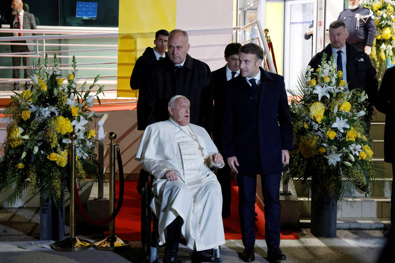 O Papa Francisco, vestido com uma túnica branca e sentado em uma cadeira de rodas, é acompanhado por um homem em um terno escuro enquanto caminha em direção a um edifício. Ao fundo, há várias pessoas, incluindo seguranças, e arranjos florais amarelos e brancos decoram a entrada. O ambiente é iluminado e há uma faixa vermelha no chão.