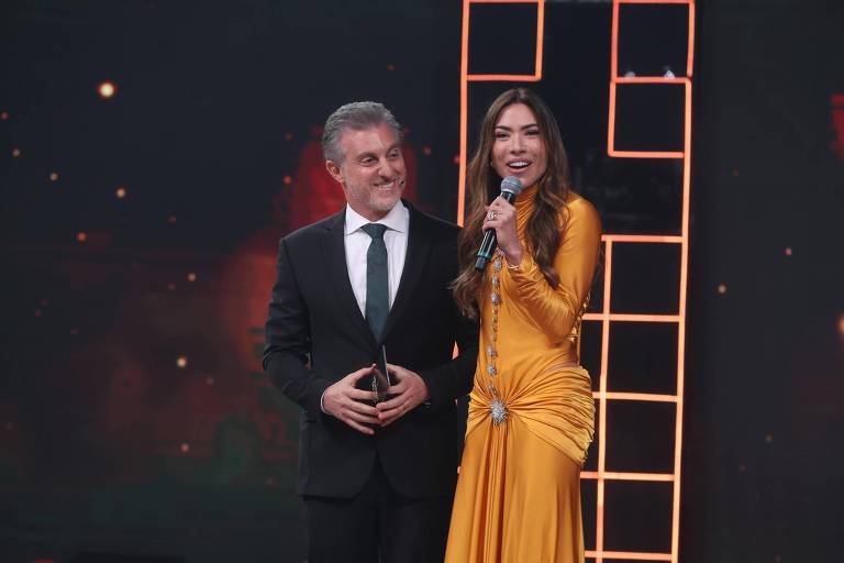 Em foto colorida, homem de terno escuro e mulher de vestido laranja aparecem juntos em cima do palco