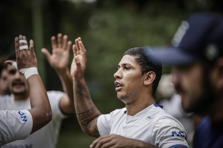 A imagem mostra um grupo de homens em um treino, com um deles levantando a mão para cumprimentar ou interagir com os outros. Eles estão vestidos com camisetas brancas e há um ambiente natural ao fundo, com árvores e vegetação. O foco principal está no homem à frente, que tem cabelo curto e uma tatuagem visível no braço.