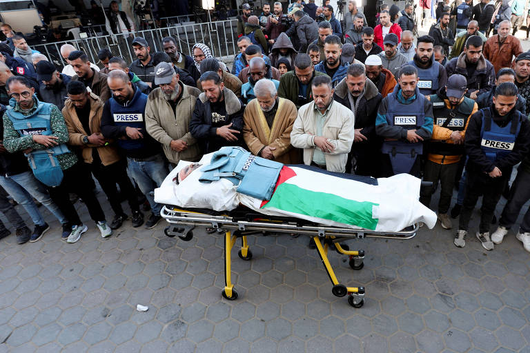 A imagem mostra um grupo de homens em um funeral, reunidos em torno de um corpo coberto por um lençol. O corpo está em uma maca, e há uma bandeira da Palestina sobre ele. Os homens estão em posição de oração, com expressões sérias, e há uma multidão ao fundo, em um ambiente urbano.