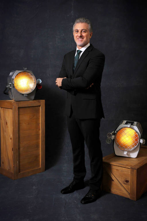 Um homem em traje formal, usando um terno preto e gravata, está posando com os braços cruzados. Ele tem cabelo grisalho e um sorriso. Ao seu lado, há dois refletores grandes, um à sua esquerda e outro à sua direita, ambos com luzes acesas, sobre caixas de madeira. O fundo é escuro e texturizado.