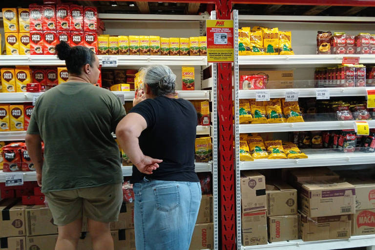 Duas mulheres estão em um corredor de supermercado， observando prateleiras cheias de produtos. A prateleira à esquerda contém pacotes de café e outros itens， enquanto a prateleira à direita exibe produtos como farinha e biscoitos. As mulheres estão de costas， uma vestindo uma camiseta verde e shorts， e a outra uma camiseta preta e jeans claros.