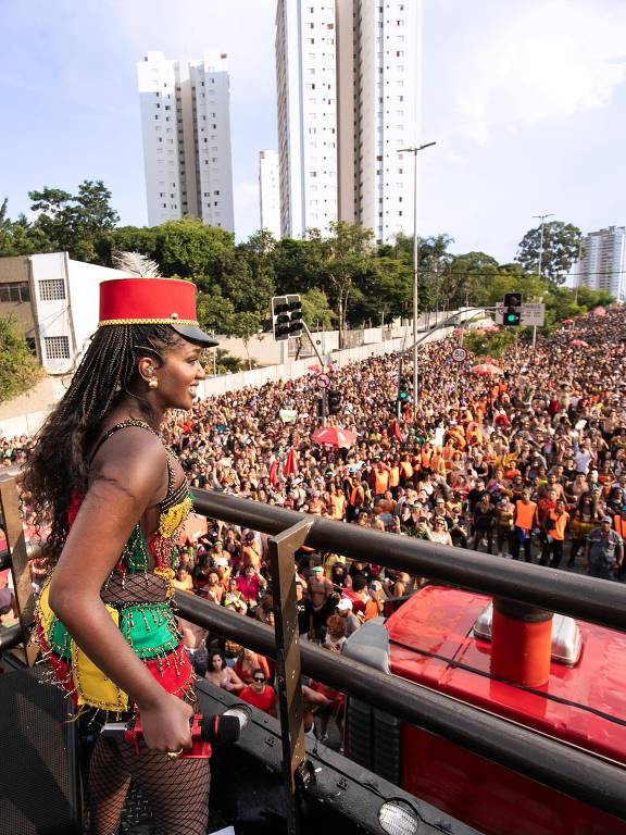 A imagem mostra uma mulher em um carro de som durante um evento de carnaval, vestindo uma fantasia colorida com detalhes em verde, amarelo e vermelho. Ela segura um microfone e observa uma multidão de pessoas abaixo, que se estende por uma rua, com muitos participantes usando roupas vibrantes. Ao fundo, há prédios altos e árvores, e a atmosfera é festiva.