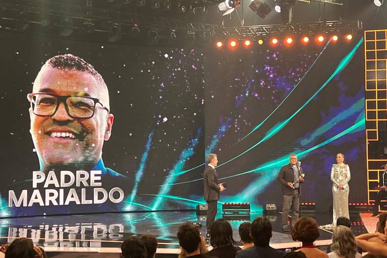 A imagem mostra um evento de premiação onde Padre Marialdo é o destaque. Ele aparece em um grande telão ao fundo, sorrindo e usando óculos, com o nome 'PADRE MARIALDO' em letras grandes. No palco, três pessoas estão presentes: um homem de terno, que parece estar apresentando, e dois outros, um deles segurando um troféu. O ambiente é iluminado com luzes coloridas e um fundo com efeitos gráficos.