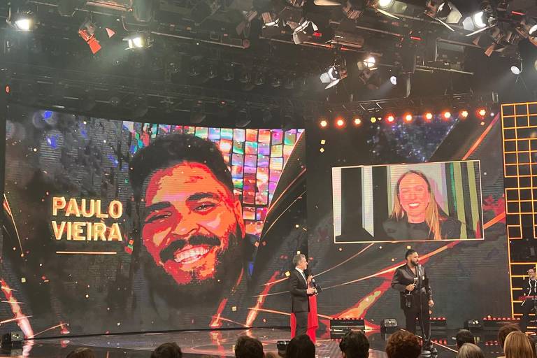 A imagem mostra um palco de evento com uma grande tela ao fundo exibindo um retrato de Paulo Vieira, que está sorrindo. O nome 'PAULO VIEIRA' está escrito em letras grandes e douradas. À frente, dois apresentadores estão conversando, um deles está vestido com um terno escuro e o outro com uma roupa vermelha. No canto superior direito, há uma tela menor mostrando uma mulher sorrindo. O ambiente é iluminado com luzes brilhantes e há uma plateia visível ao fundo.