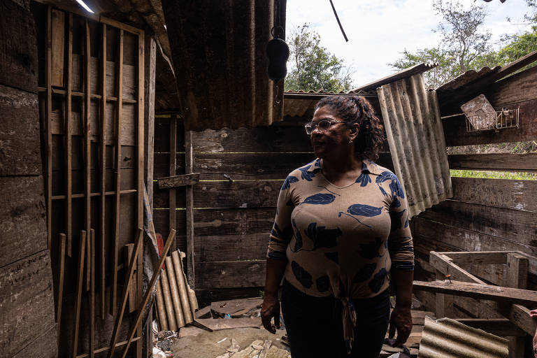 A imagem mostra uma mulher em um ambiente interno rústico, com paredes de madeira e algumas partes expostas. Ela está de pé, olhando para o lado, vestindo uma blusa de mangas longas. O espaço ao redor parece desorganizado, com tábuas de madeira e outros materiais espalhados pelo chão