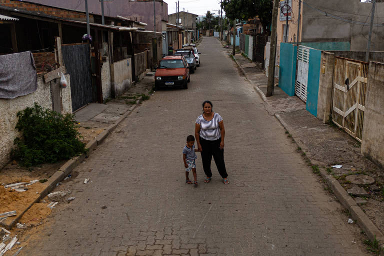 A imagem mostra uma rua residencial com casas de um lado e um carro estacionado. No centro da rua, uma mulher está de pé ao lado de uma criança. A mulher usa uma camiseta branca e calças escuras, enquanto a criança está vestida com uma camiseta cinza e calças escuras