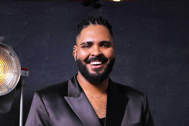 Um homem sorridente está posando em um fundo escuro, vestindo um traje formal preto brilhante. Ele segura um troféu em uma das mãos e está apoiado em uma caixa de madeira com uma perna levantada. Ao lado dele, há um holofote que emite uma luz amarela. O homem tem cabelo trançado e uma barba bem cuidada.