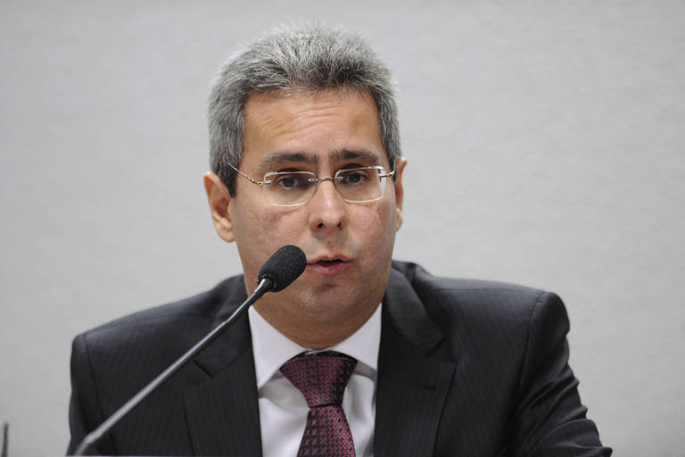 Um homem com cabelo grisalho e óculos está falando em um microfone durante uma audiência. Ele usa um terno escuro e uma gravata vinho, e parece estar em um ambiente formal, possivelmente uma sala de reuniões ou uma comissão.