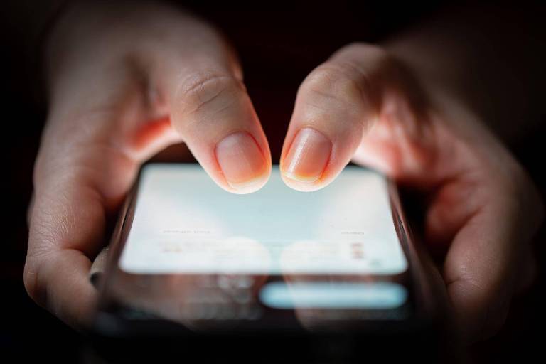 A imagem mostra duas mãos segurando um smartphone. As pontas dos dedos estão tocando a tela do dispositivo, que emite uma luz suave. O fundo é escuro, destacando as mãos e o telefone.