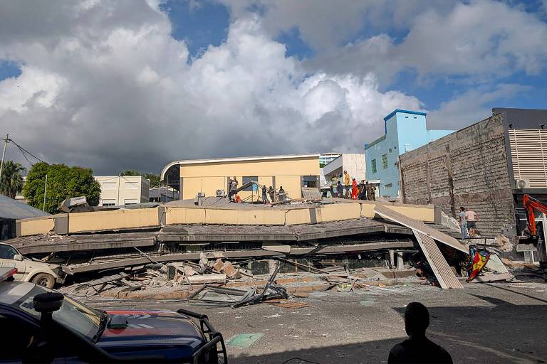 A imagem mostra um edifício que desabou, com escombros visíveis no chão. Algumas pessoas estão em cima da estrutura colapsada, enquanto outras observam ao fundo. O céu está parcialmente nublado, e há prédios ao redor, incluindo um de cor azul. A cena parece ser de um local urbano afetado por um desastre.