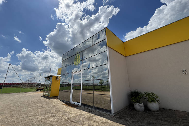 A imagem mostra a fachada de um edifício moderno com paredes de vidro refletindo o céu azul e nuvens brancas. O edifício possui detalhes em amarelo e uma porta de vidro. À esquerda, há uma pequena estrutura amarela com uma janela. Ao fundo, é possível ver um campo de futebol cercado por uma rede. O chão é pavimentado com pedras e há vasos com plantas na parte inferior direita.