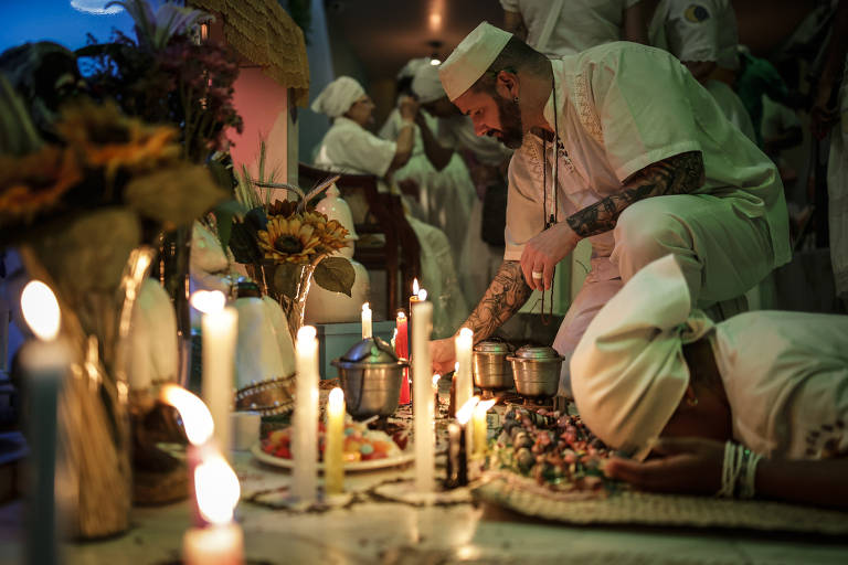 A imagem mostra um ritual religioso em um ambiente com iluminação suave. Um homem, vestido com roupas brancas e um chapéu, está ajoelhado em frente a um altar, onde há velas acesas, flores e oferendas. Ao fundo, outras pessoas também participam do ritual, vestindo roupas brancas. O ambiente é decorado com elementos que sugerem uma cerimônia espiritual.