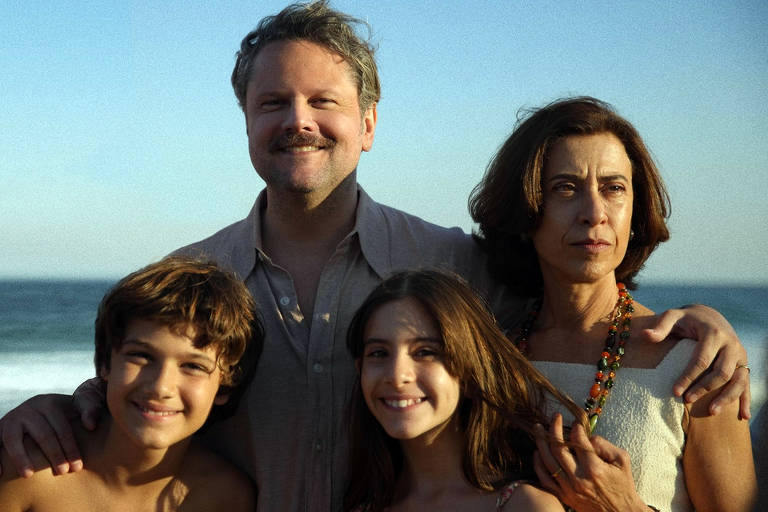 A imagem mostra uma família posando na praia. No centro， um homem com bigode e sorriso， vestindo uma camisa clara， está abraçado a uma mulher com cabelo castanho e uma blusa clara. À esquerda， um menino com cabelo castanho e curto， sorrindo， e à direita， uma menina com cabelo longo e castanho， também sorrindo. O fundo apresenta o mar e um céu claro.