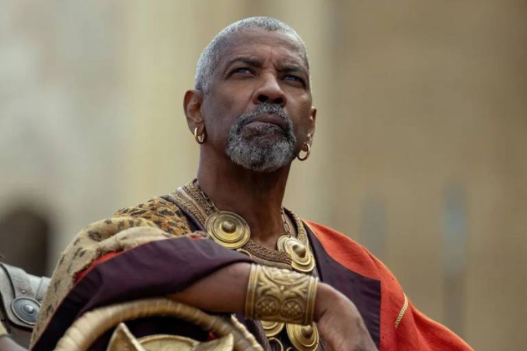 A imagem mostra um homem com cabelo grisalho e barba, usando um traje histórico que inclui colares e um manto vermelho. Ele está olhando para cima, com uma expressão séria, e possui um brinco em uma das orelhas. O fundo é desfocado, sugerindo um ambiente ao ar livre.