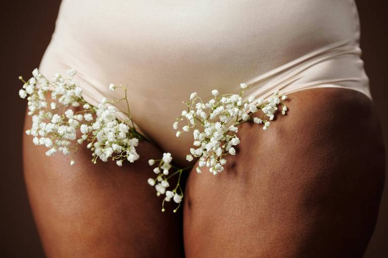 A imagem mostra a parte inferior do corpo de uma pessoa， com um fundo neutro. A pessoa está usando uma calcinha de cor clara e tem ramos de flores brancas， possivelmente gypsophila， posicionados em cada lado das coxas.