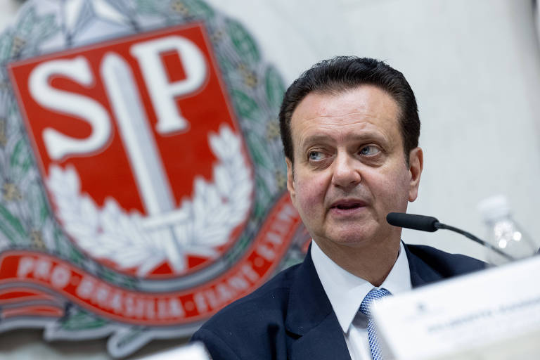 A imagem mostra Gilberto Kassab, homem branco de terno escuro e gravata, sentado em uma mesa, falando. Ele tem cabelo escuro e está olhando para o lado. Ao fundo, há um grande logotipo da Assembleia Legislativa de SP, que é vermelho e branco, com uma espada e folhas ao redor. A mesa tem uma garrafa de água e um documento visível.