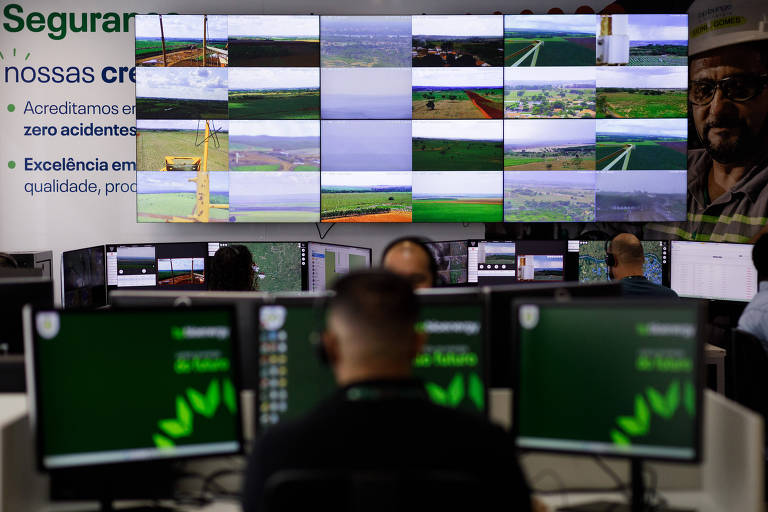 A imagem mostra um centro de monitoramento com várias telas exibindo imagens de diferentes áreas. No primeiro plano， há pessoas sentadas em frente a computadores com telas verdes. Ao fundo， uma grande tela exibe múltiplas imagens， algumas com paisagens e outras com informações visuais. Há também um banner na parte superior com texto em português.