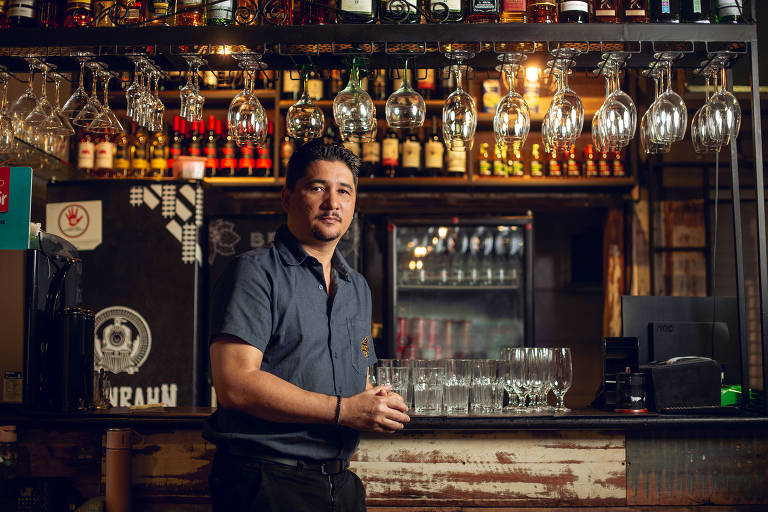 Um homem está posicionado em frente a um balcão de bar， com prateleiras cheias de garrafas de bebidas e copos pendurados acima dele. Ele está usando uma camisa de botão escura e tem uma expressão amigável. Ao fundo， há uma geladeira com bebidas e uma decoração rústica.