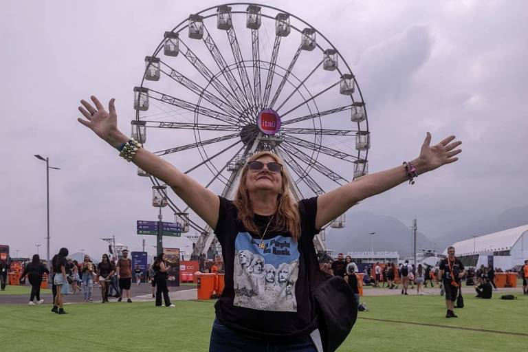 Uma mulher está em primeiro plano， com os braços abertos e sorrindo， em um ambiente de festival. Ao fundo， há uma roda-gigante grande e várias pessoas caminhando. O céu está nublado.