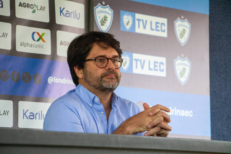 Um homem com cabelo escuro e barba está sentado em uma mesa durante uma coletiva de imprensa. Ele usa óculos e uma camisa azul clara. Ao fundo， há um painel com logotipos do Londrina EC e patrocinadores， incluindo 039;TV LEC039; e 039;AGROPLAY039;.