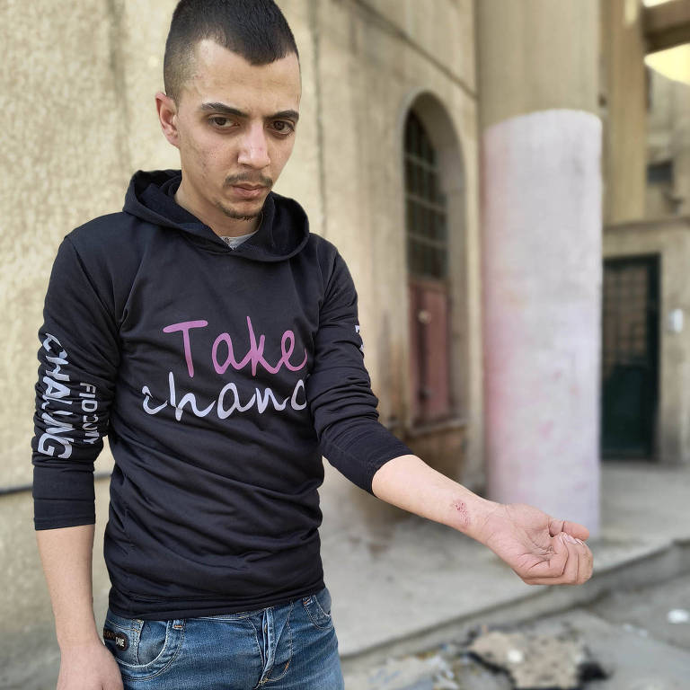 Um jovem está em pé em uma rua, vestindo uma camiseta preta com a frase 'Take the Risk' em letras coloridas. Ele usa jeans e tem cabelo curto. O fundo mostra uma parede de edifício e um chão de pedra, com alguns detritos visíveis