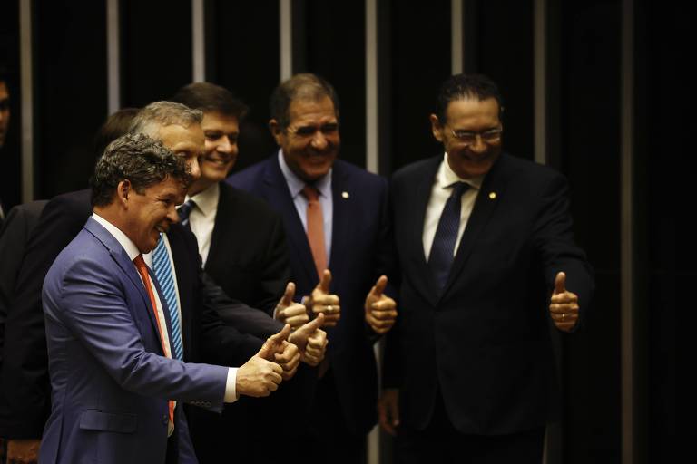 Um grupo de cinco homens está posando para a foto em um ambiente formal. Eles estão sorrindo e fazendo sinal de positivo com os polegares. Os homens estão vestidos com trajes formais, incluindo paletós e gravatas. O fundo é escuro, com linhas verticais que sugerem um ambiente institucional.