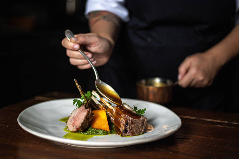 A imagem mostra um prato gourmet sendo servido. Um chef， com uma camisa branca e avental escuro， está usando uma colher para adicionar um molho sobre a carne e acompanhamentos no prato. O prato contém pedaços de carne， abóbora e um purê verde， decorado com uma folhinha de erva. Ao fundo， há um recipiente metálico.