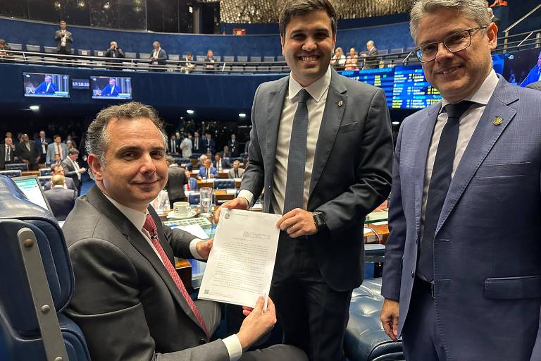 Três homens estão em um ambiente de plenário do Senado. Um deles， à esquerda， está sentado e segurando um documento， enquanto os outros dois estão em pé ao seu lado， sorrindo. O ambiente é bem iluminado， com cadeiras azuis e telas ao fundo mostrando informações. Todos estão vestidos formalmente.