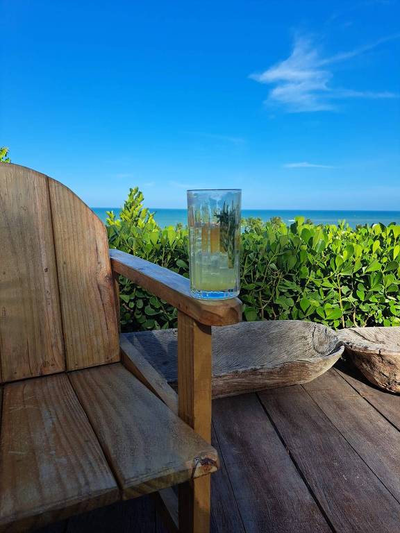 cadeira de madeira com mar ao fundo e um drinque apoiado no braço