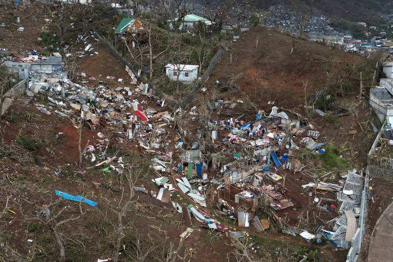 A imagem mostra uma área devastada por um desastre natural, com destroços de casas e árvores danificadas. O solo está coberto de escombros, incluindo telhas e partes de estruturas. Ao fundo, é possível ver uma área mais densa de vegetação e algumas construções intactas.