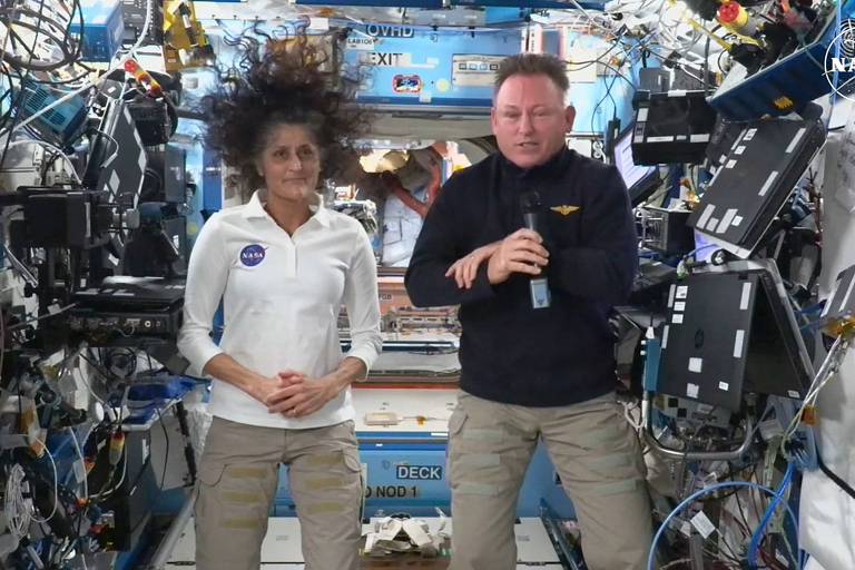 A imagem mostra dois astronautas dentro da Estação Espacial Internacional. Um astronauta está à esquerda， vestindo uma camisa branca com um emblema， e o outro à direita， usando uma camisa preta. O ambiente ao redor é repleto de equipamentos e cabos， característico do interior da estação espacial.