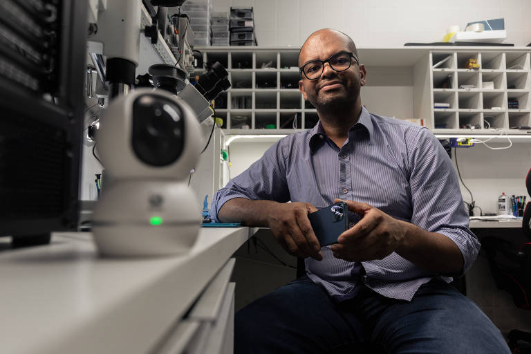 Um homem sentado em um laboratório， segurando um dispositivo móvel. Ele está usando uma camisa de botão e óculos. À sua esquerda， há uma câmera de segurança e ao fundo， prateleiras com diversos objetos organizados. A iluminação é suave， destacando o ambiente de trabalho.