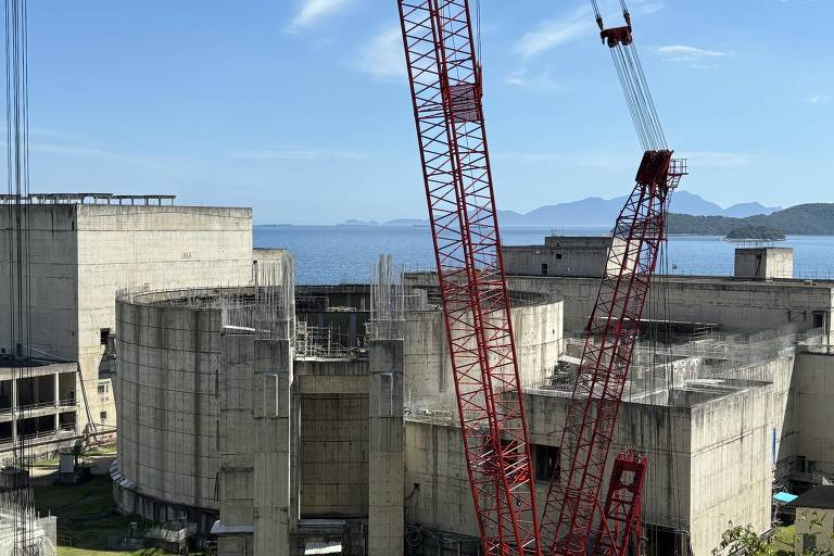 A imagem mostra uma usina com estruturas de concreto， incluindo tanques e edifícios. Há uma grua vermelha em operação， e ao fundo é possível ver um corpo d039;água e montanhas ao longe sob um céu claro.
