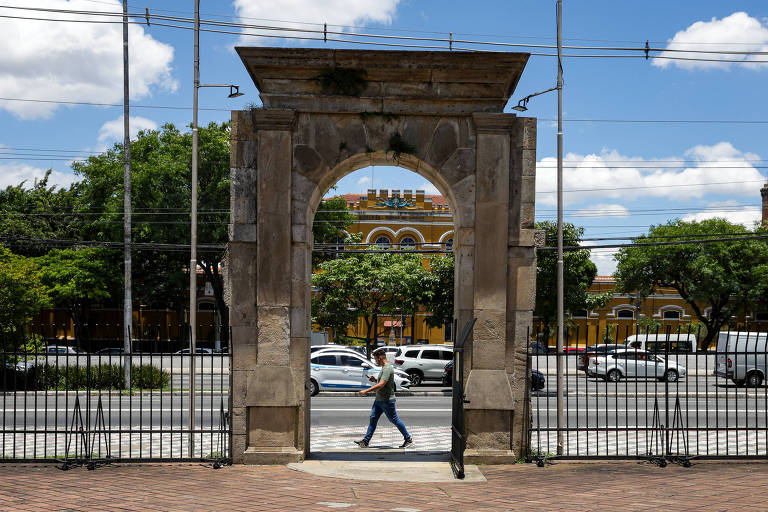 Homem caminha na frente de um arco grande, no meio da rua