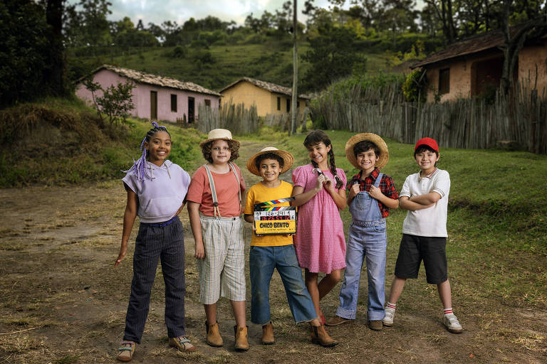 Seis crianças estão posando em um ambiente rural， com casas de cores pastel ao fundo. Elas estão vestidas de maneira casual， algumas usando chapéus. A criança no centro segura uma placa de filmagem. O cenário é verde， com árvores e um céu nublado