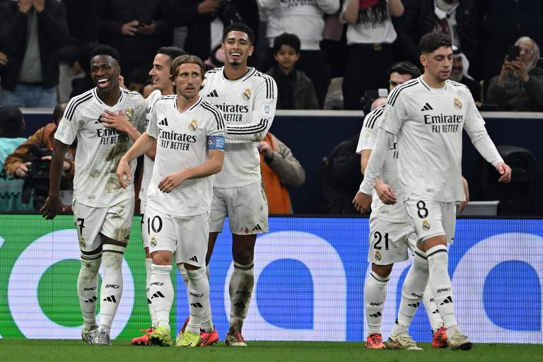 Um grupo de jogadores do Real Madrid， vestidos com uniformes brancos， celebra em campo. Eles estão sorrindo e interagindo entre si， com um jogador no centro recebendo os cumprimentos dos colegas. O fundo mostra uma torcida animada.
