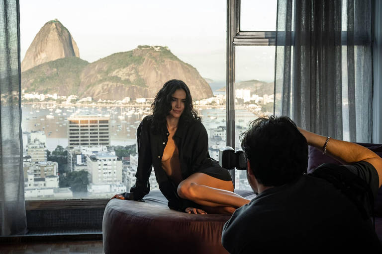 A imagem mostra uma mulher com cabelo cacheado, vestindo uma camisa preta, posando em um sofá enquanto um fotógrafo a captura com uma câmera. Ao fundo, há uma vista panorâmica da cidade, incluindo o Pão de Açúcar e um mar com barcos. A luz natural entra pela janela, iluminando a cena.