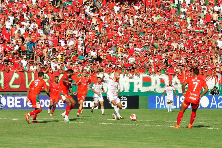 A imagem mostra um jogo de futebol em um estádio lotado. A torcida， predominantemente vestida de vermelho， está animada e aplaudindo. No campo， jogadores de duas equipes estão em ação， com um jogador em destaque， vestindo uma camisa branca， prestes a chutar a bola. Ao fundo， há uma grande faixa verde e vermelha com a palavra 039;TORCIDA039;.