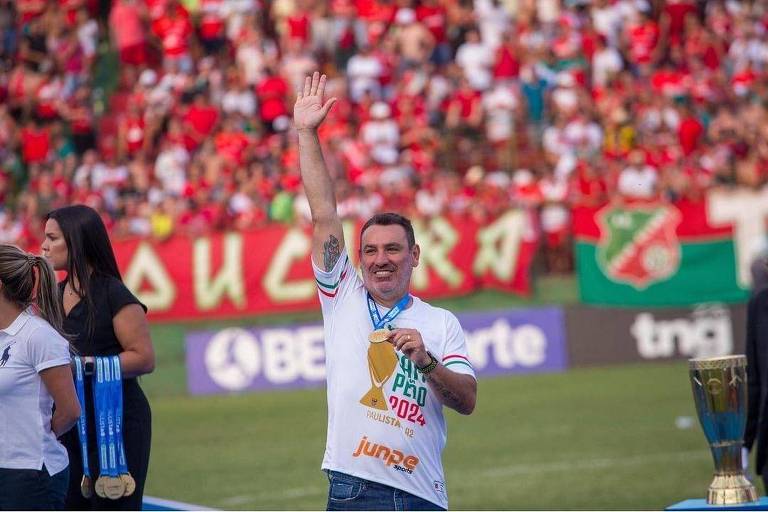 Um homem com uma camiseta branca e medalha de campeão levanta a mão em um gesto de celebração. Ele está em um campo de futebol， cercado por uma multidão de torcedores vestidos em sua maioria de vermelho. Ao fundo， há uma faixa com o nome do time e um troféu ao lado dele. Duas mulheres estão próximas， uma vestindo uma camiseta branca e a outra com uma blusa preta.