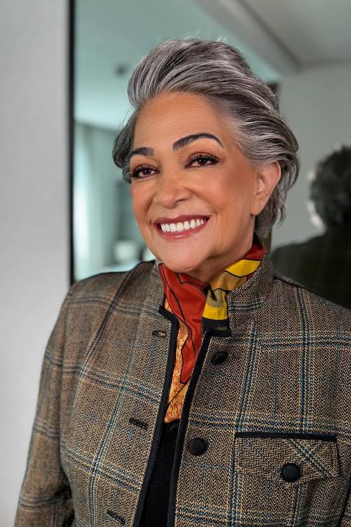 A imagem mostra uma mulher sorridente com cabelo grisalho e estilizado. Ela está usando um casaco xadrez com detalhes em preto e uma blusa preta por baixo. Um lenço colorido, predominantemente vermelho e amarelo, está amarrado ao redor do pescoço. O fundo é desfocado, sugerindo um ambiente interno, possivelmente uma sala de estar ou um quarto.