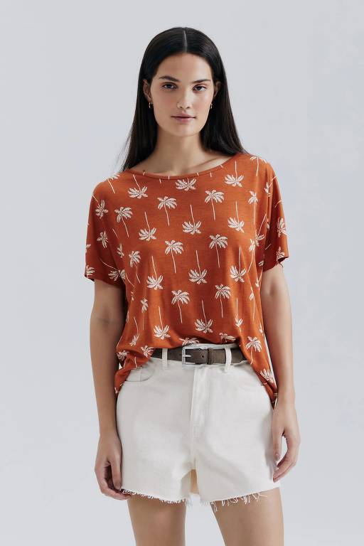 Uma jovem mulher está posando em pé, usando uma camiseta de cor laranja com estampas de animais em tons mais claros. Ela está vestindo um short branco e um cinto escuro. O fundo da imagem é neutro, destacando a modelo e suas roupas.