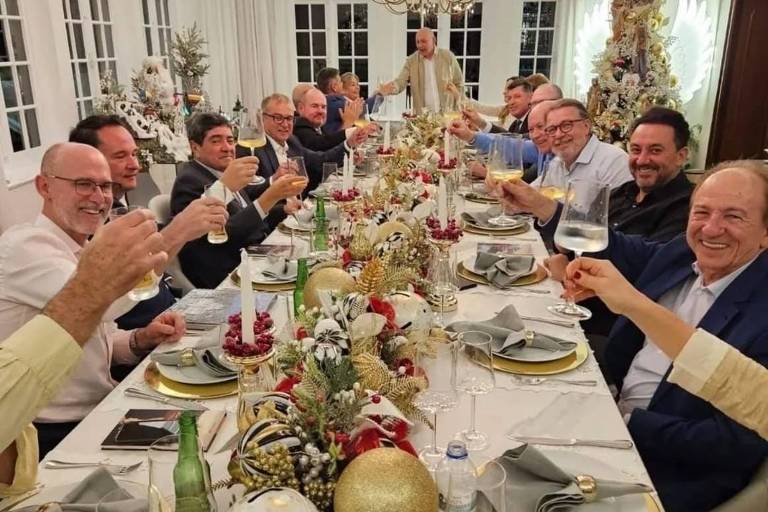 A imagem mostra um grupo de pessoas， a maioria homens， reunidos em torno de uma longa mesa de jantar decorada com flores， velas e enfeites natalinos. Todos estão levantando seus copos em um brinde， sorrindo e parecendo se divertir. O ambiente é iluminado por um grande lustre e há janelas ao fundo， sugerindo um ambiente festivo e acolhedor.