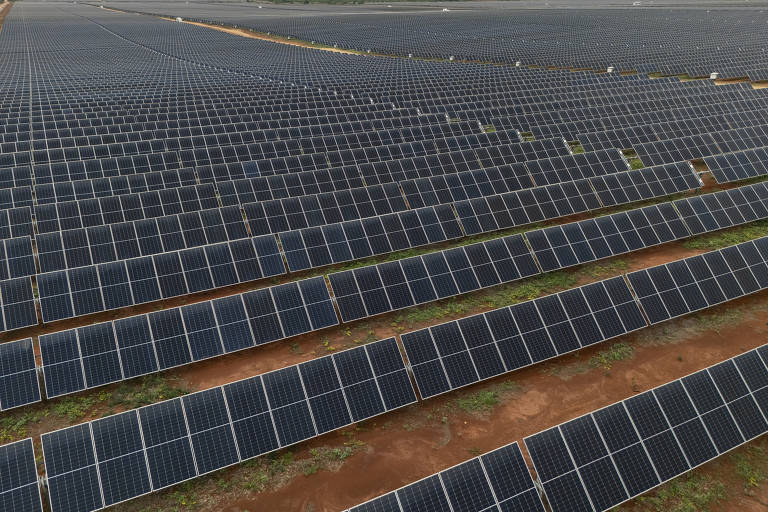 A imagem mostra um grande campo de painéis solares dispostos em linhas organizadas. Os painéis são predominantemente pretos e estão alinhados em várias fileiras， cobrindo uma vasta área de terreno. O céu está nublado， e há algumas pessoas trabalhando entre os painéis.
