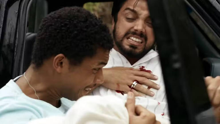 A imagem mostra duas pessoas dentro de um carro. Um homem com cabelo crespo e pele clara está segurando um homem com cabelo escuro e pele morena, que parece estar ferido, com sangue visível em sua camisa branca. O homem ferido está com uma expressão de dor, enquanto o outro parece angustiado. O ambiente é interno de um carro, com janelas e um fundo desfocado.