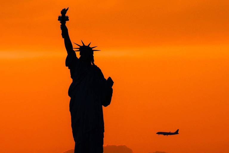 A imagem mostra a silhueta da Estátua da Liberdade contra um céu laranja ao pôr do sol, com um avião voando ao fundo