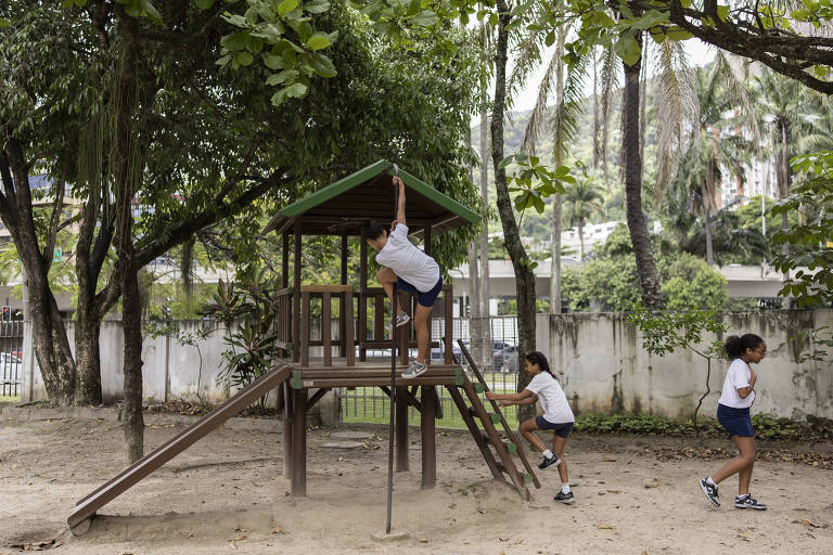 A imagem mostra um parque infantil com uma estrutura de madeira. Uma criança está subindo na parte superior da estrutura， enquanto outras duas crianças estão brincando ao redor. O ambiente é cercado por árvores e há um chão de areia sob a estrutura. O dia parece nublado.