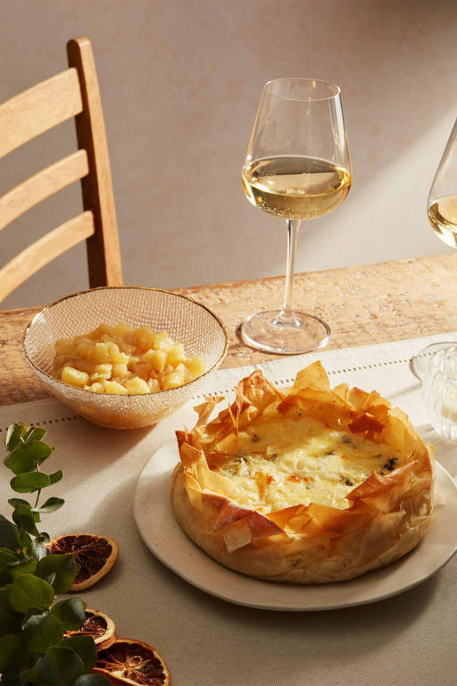 A foto mostra uma mesa com um gateau de batatas envolto em massa fina e dourada. Ao fundo， um travessa com frutas e duas taças de vinho branco