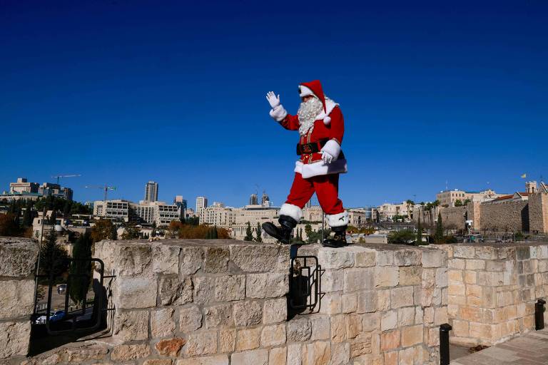 Um homem vestido de Papai Noel, com um traje vermelho e branco, está em cima de uma muralha de pedra, acenando com a mão. Ao fundo, há uma vista da cidade de Jerusalém sob um céu azul claro.