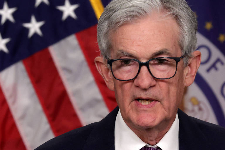 Imagem mostra Jerome Powell， um homem com cabelo grisalho e óculos que está falando em frente a uma bandeira dos Estados Unidos. O fundo apresenta estrelas brancas em um campo azul e listras vermelhas e brancas.